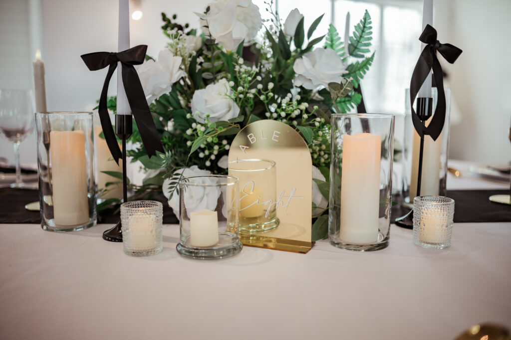 Elegant wedding tablescape featuring white roses, lush greenery, black ribbon accents, golden table markers, and glowing candles in glass holders for a classic and moody ambiance.